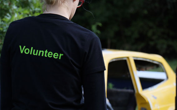 Grünenthal employee standing outside in front of a broken car wearing a black sweater with green print “Volunteer”on the back.